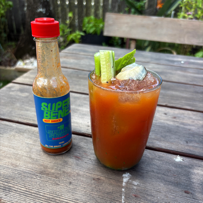 Glass of Bloody Mary with celery and oyster, next to a bottle of Super Benne hot sauce on a wooden table.