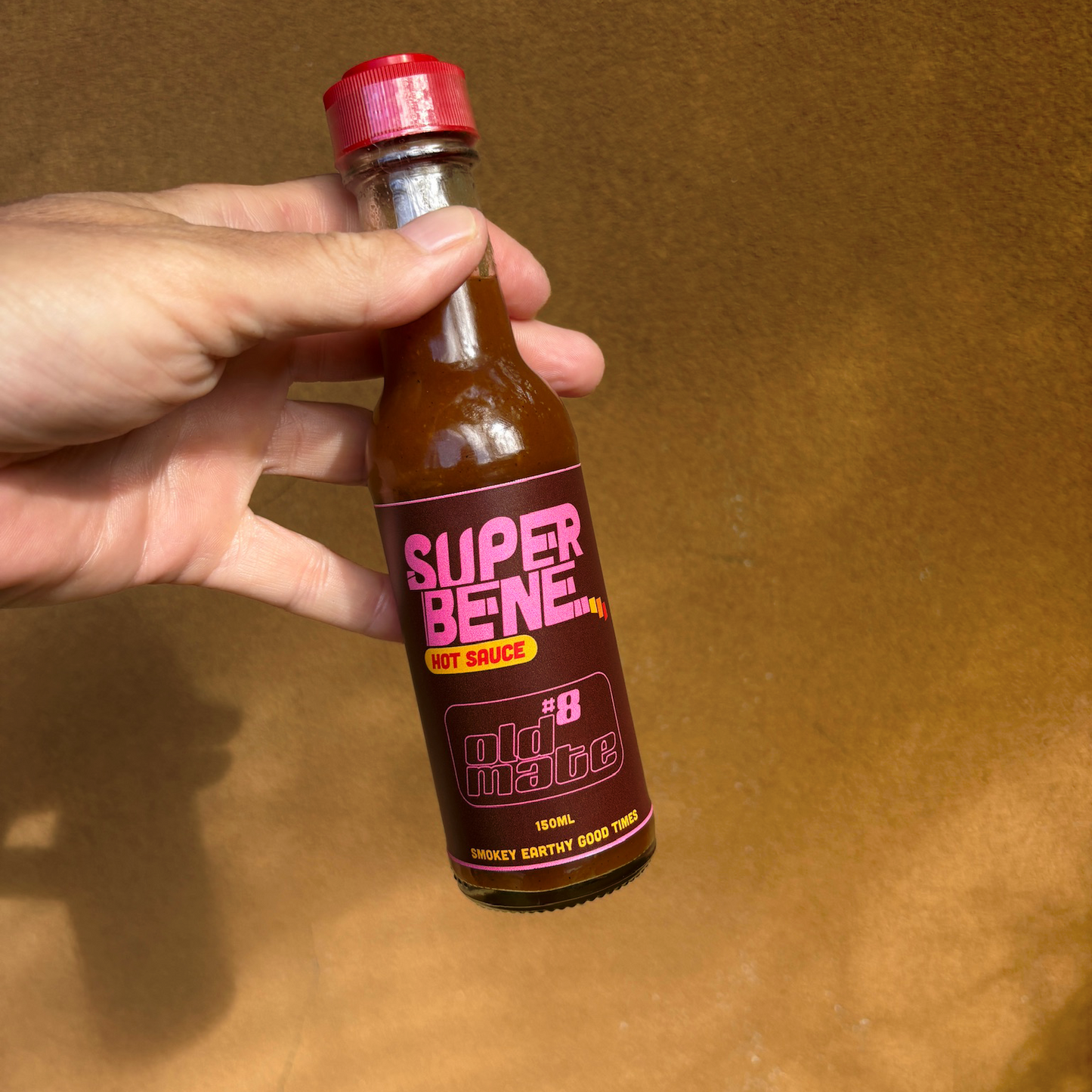 Hand holding a bottle of Super Bene hot sauce on a brown background