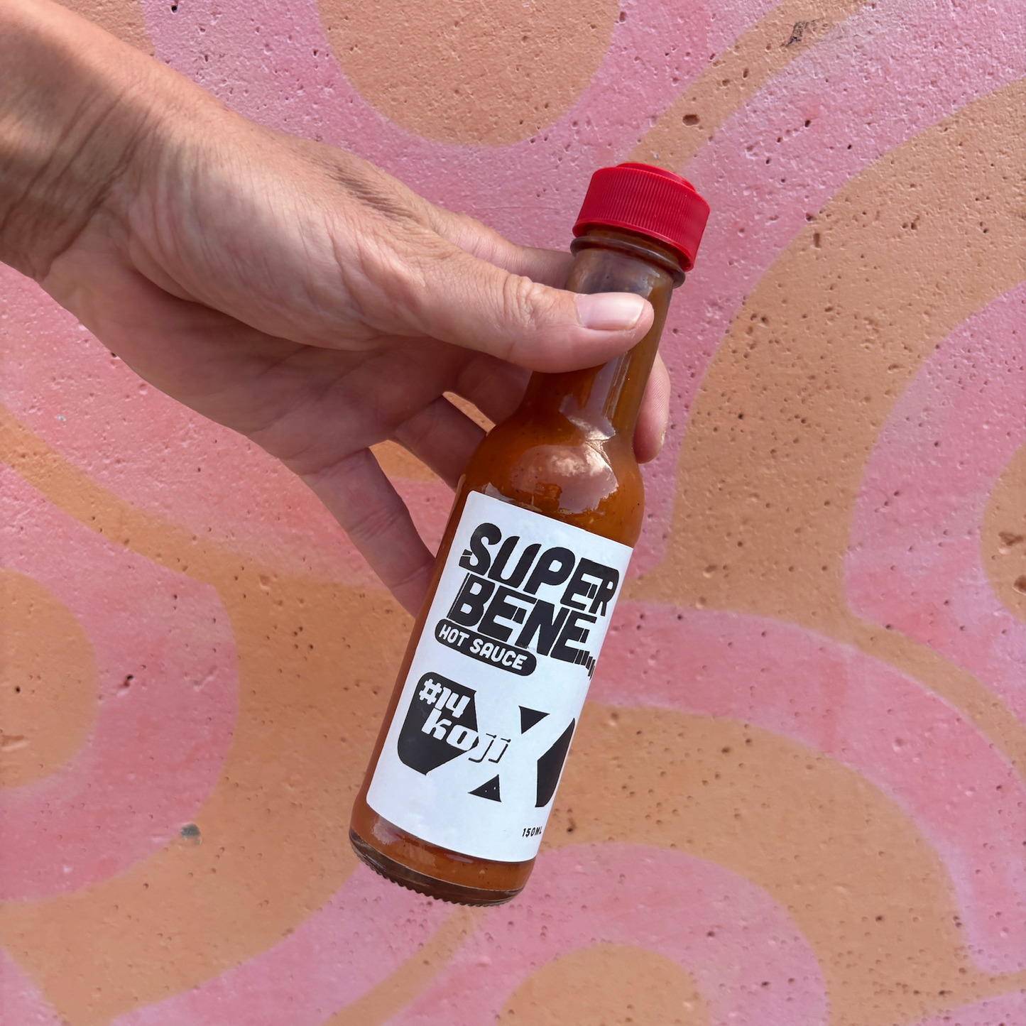 Hand holding a bottle of Super Bene hot sauce against a pink and brown abstract background