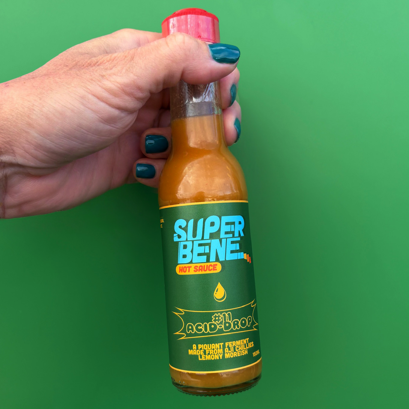 Hand holding a bottle of Super Benne hot sauce against a green background