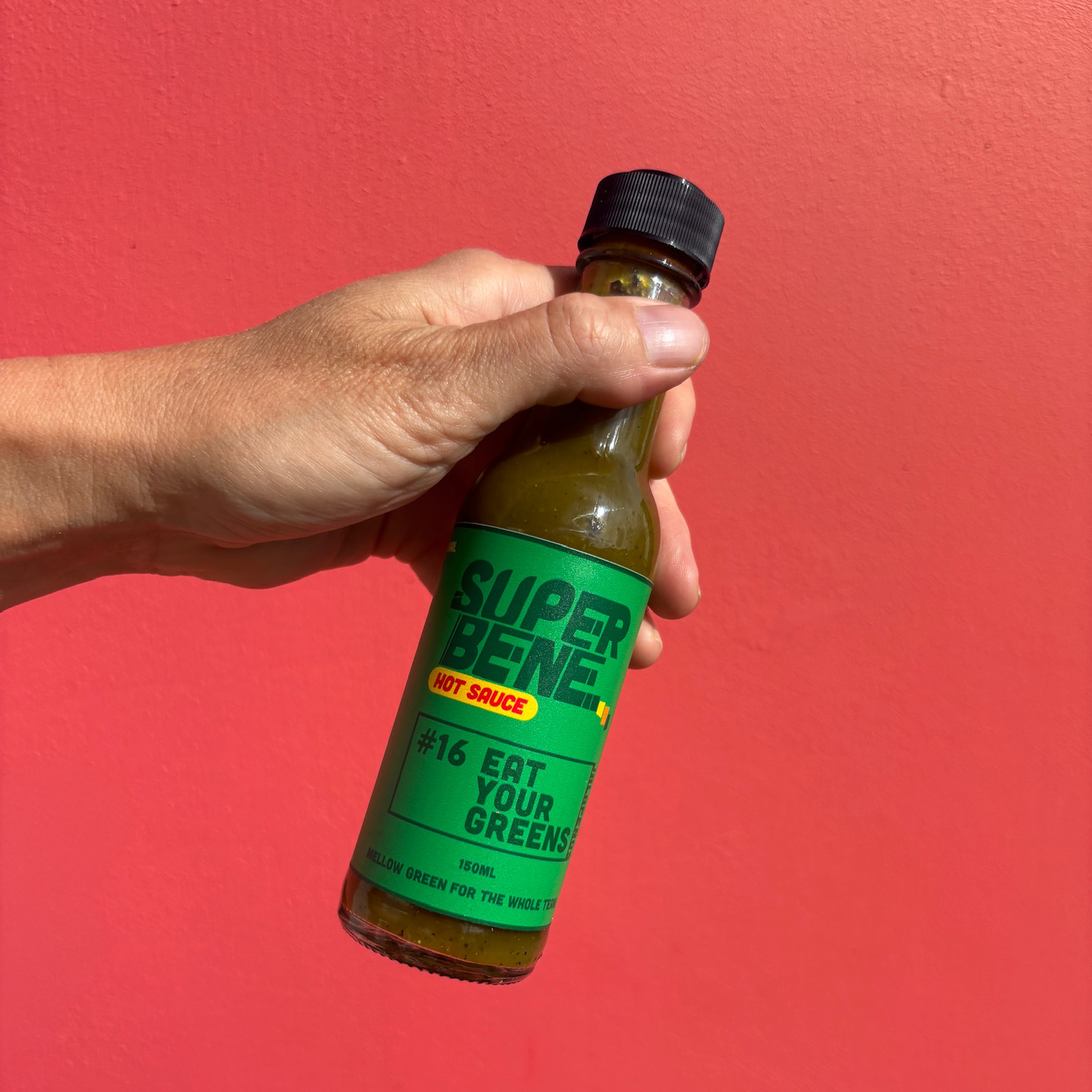 Hand holding a bottle of Super Bene hot sauce against a red background