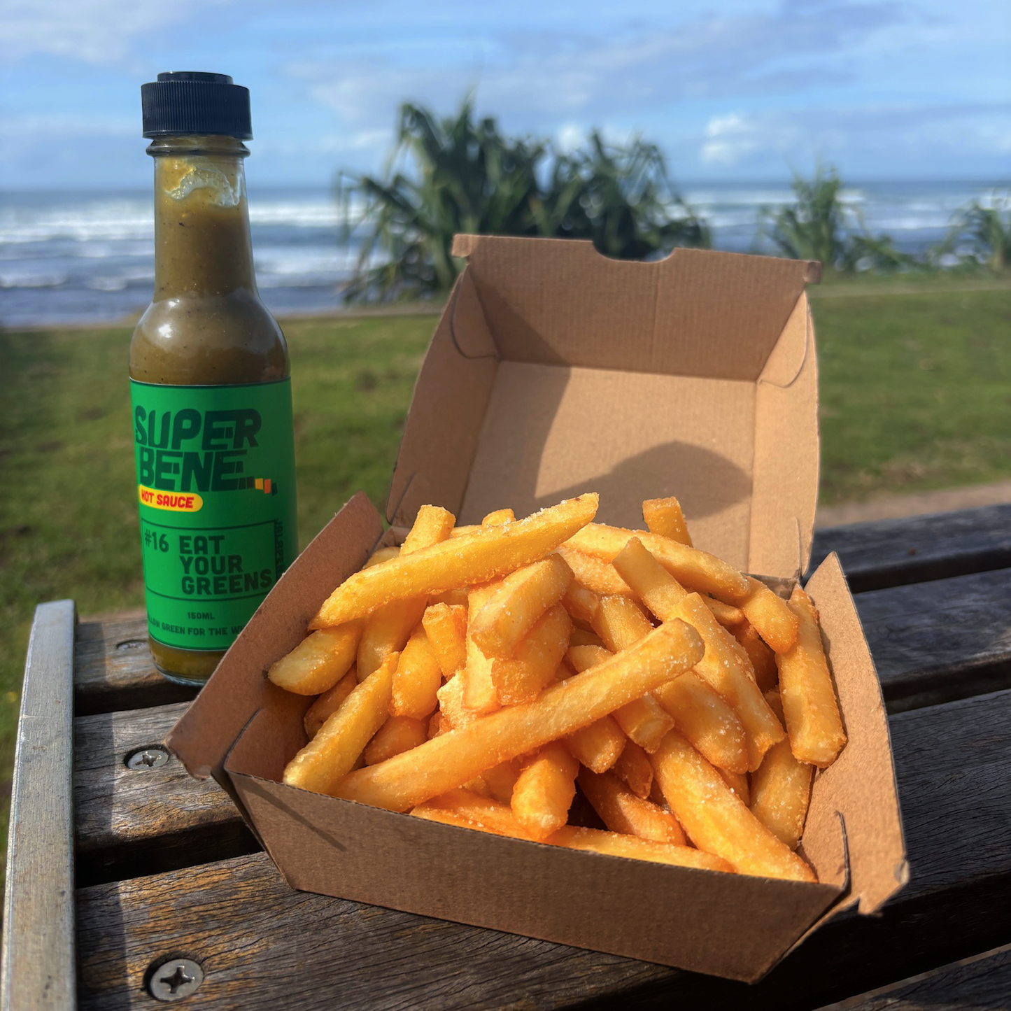 Box of fries and bottle of Super Bene sauce on a wooden surface with a beach background