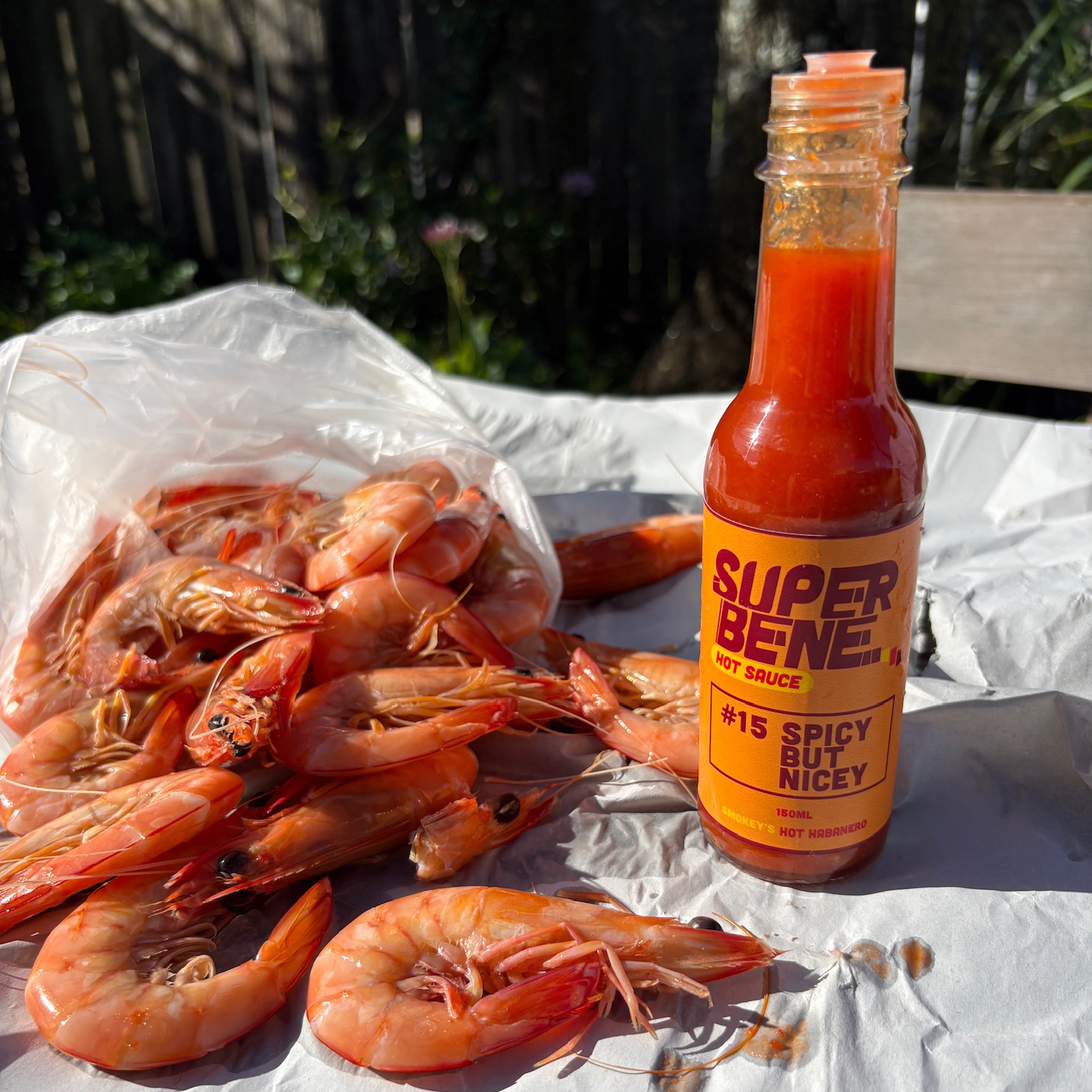 Bottle of Super Bene hot sauce with a bag of prawns on a white surface outdoors.