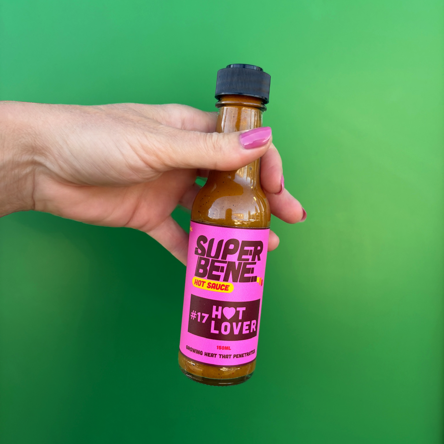 Hand holding a bottle of Super Bene hot sauce against a green background hot lover