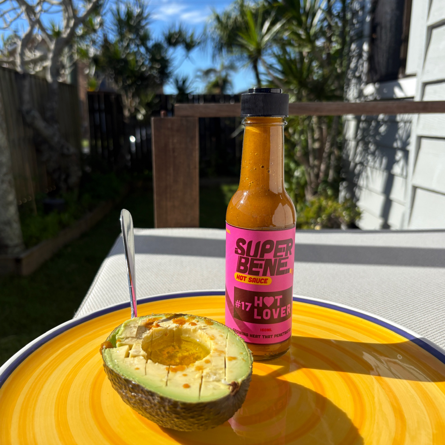 Super Bene hot sauce bottle next to a halved avocado on a yellow plate outdoors.