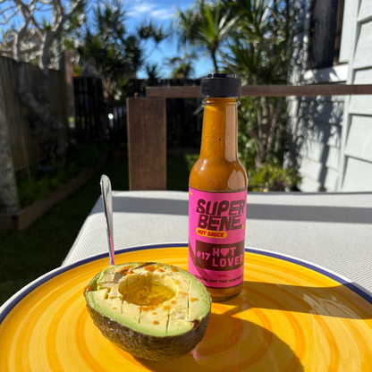 Super Bene hot sauce bottle next to a halved avocado on a yellow plate outdoors.